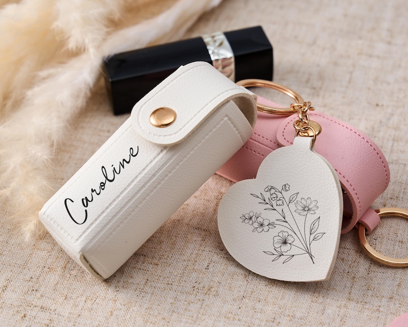 May include: A white lipstick case with a gold snap closure and the name Caroline printed in black script. Attached to the case is a heart-shaped white leather keychain featuring a black line drawing of a floral bouquet. A pink case is nearby.