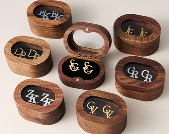 May include: A collection of personalized initial earrings displayed in oval wooden boxes with clear lids. The earrings are gold-colored, and the initials are visible through the lids. The boxes are arranged on a light-colored surface.