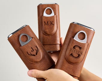 May include: Three brown leather cigar cases with silver accents and built-in cigar cutters. Each case features a different engraved design: initials "MK", a deer antler, and the name "Steven". The cases are held in a person's hands.