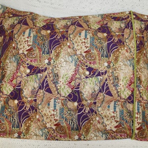 May include: Rectangular cushion with a patterned fabric in purple, beige, red, and green. The design incorporates floral and geometric shapes. A green cord trim is visible along one edge.