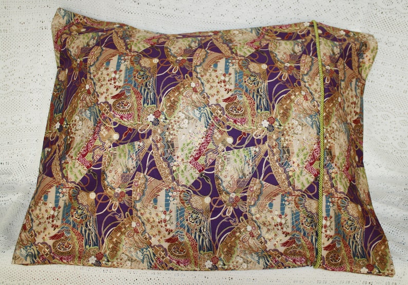 May include: Rectangular cushion with a patterned fabric in purple, beige, red, and green. The design incorporates floral and geometric shapes. A green cord trim is visible along one edge.