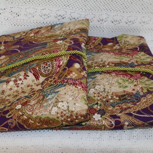 May include: Two rectangular fabric pouches with a Japanese-inspired design. The fabric features a mix of purple, gold, red, and blue hues with floral and fan motifs. A green and gold rope trim accents the edges.