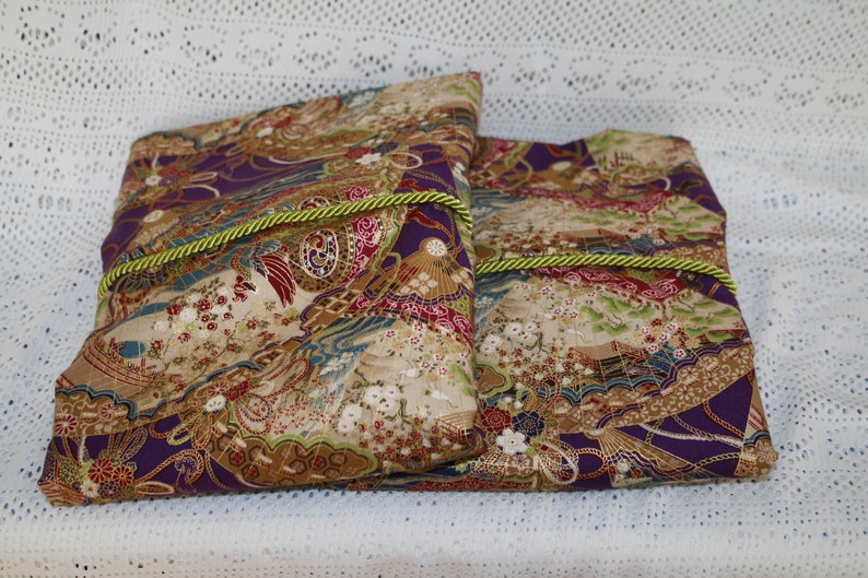 May include: Two rectangular fabric pouches with a Japanese-inspired design. The fabric features a mix of purple, gold, red, and blue hues with floral and fan motifs. A green and gold rope trim accents the edges.