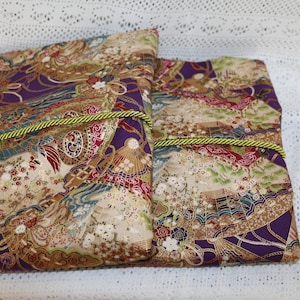 May include: Two fabric-covered books with a Japanese-inspired design. The covers feature a pattern of fans, flowers, and landscapes in shades of purple, beige, and red. A green cord wraps around each book, securing them. The books are placed on a white, lace-like surface.