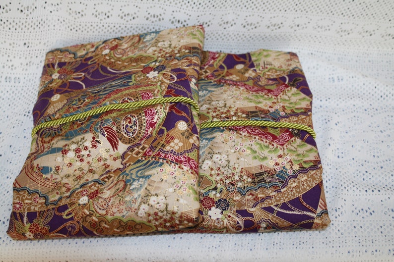 May include: Two fabric-covered books with a Japanese-inspired design. The covers feature a pattern of fans, flowers, and landscapes in shades of purple, beige, and red. A green cord wraps around each book, securing them. The books are placed on a white, lace-like surface.