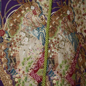 May include: Close-up of patterned fabric with a vibrant design. The fabric features purple, gold, red, and green. The design includes floral and bird motifs, with a green rope trim down the centre.