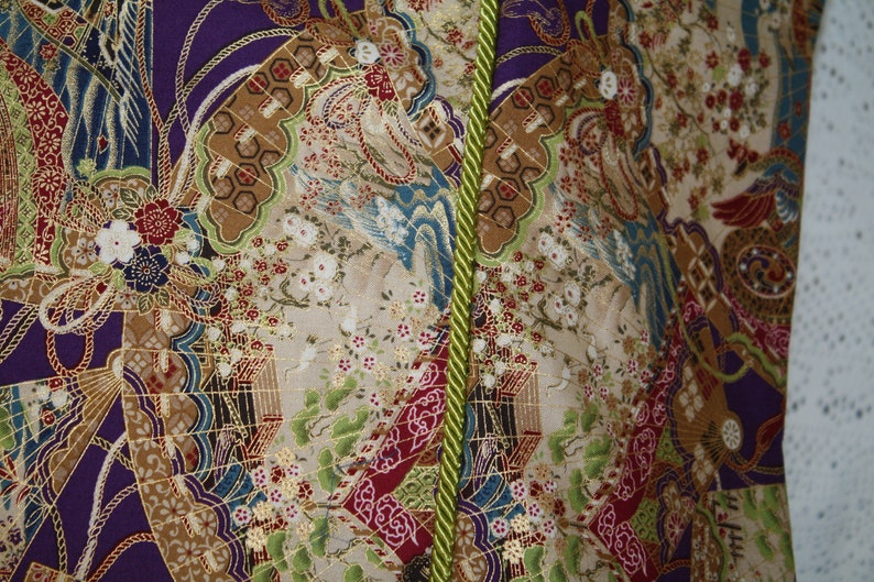 May include: Close-up of patterned fabric with a vibrant design. The fabric features purple, gold, red, and green. The design includes floral and bird motifs, with a green rope trim down the centre.