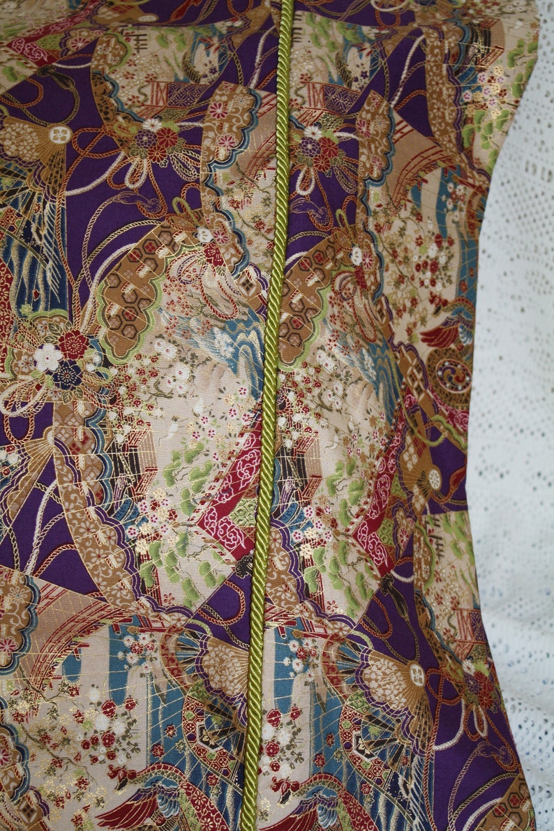 May include: Close-up of a patterned fabric with a vibrant design. The fabric features a mix of colours, including purple, beige, and green, with a repeating pattern of fans, flowers, and other decorative elements. A green cord runs vertically down the centre.