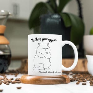 Puede incluir: Taza de cerámica blanca con un dibujo de línea negra de un gato con el texto "What, you egg?" y "Macbeth (Acto 4, Escena 1)" impreso en ella.