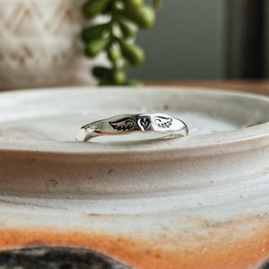 May include: A silver ring featuring a heart and wings design. The ring is resting on a ceramic dish.