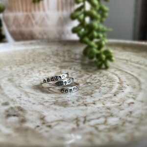 May include: Three silver rings stacked on a white ceramic surface. The top ring has the name "Hadlee" engraved on it. The middle ring has a small heart and angel wings engraved on it. The bottom ring has the name "Connor" engraved on it.