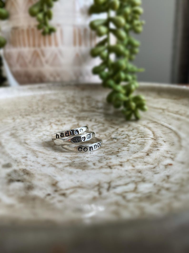 May include: Three silver rings stacked on a white ceramic surface. The top ring has the name "Hadlee" engraved on it. The middle ring has a small heart and angel wings engraved on it. The bottom ring has the name "Connor" engraved on it.