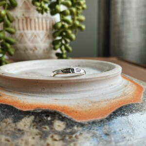 May include: A silver ring featuring a heart and wings design. The ring is resting on a ceramic dish with a brown and white glaze.