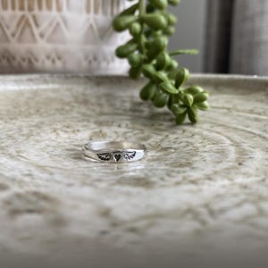 May include: A silver ring featuring a heart and wings design. The ring is displayed on a light brown surface.