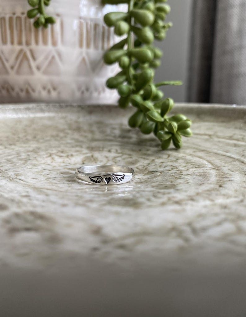 May include: A silver ring featuring a heart and wings design. The ring is displayed on a light brown surface.