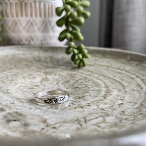May include: A silver ring featuring a heart and wings design. The ring is displayed on a light brown textured surface.