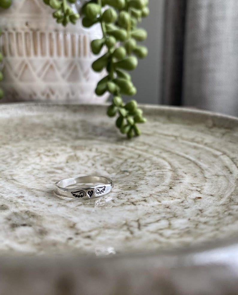 May include: A silver ring featuring a heart and wings design. The ring is displayed on a light brown textured surface.