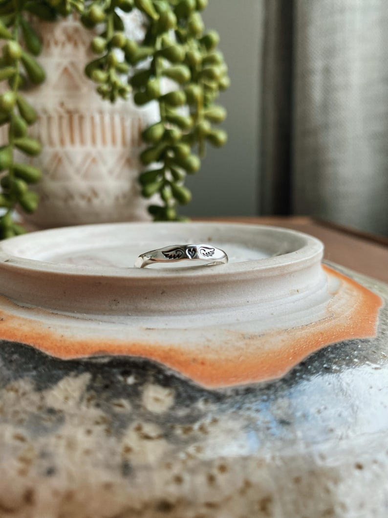 May include: A silver ring featuring a delicate design of angel wings. The ring is simple and elegant, ideal for everyday wear.