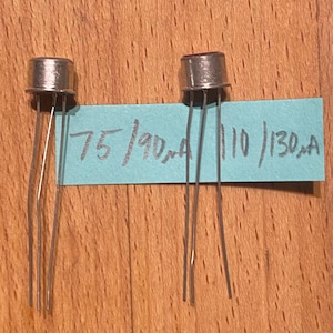 May include: Two silver-colored metal electronic transistors with three thin wire leads each. A light blue rectangular paper strip is positioned behind the components with the handwritten text 75/90µA and 110/130µA in black ink.