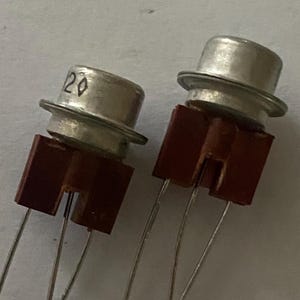 Może przedstawiać: Dwa srebrne tranzystory z brązowymi plastikowymi podstawami i trzema wyprowadzeniami każdy. Tranzystor po lewej stronie ma nadrukowaną na górze liczbę "20".