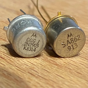 May include: Two silver transistors with gold accents. The transistor on the left has the text "A CGG J 2N2905 K8314" printed on it. The transistor on the right has the text "M 5A862 913" printed on it.