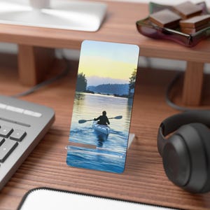 May include: A phone case with a scenic image of a person kayaking on a lake at sunset. The lake is surrounded by trees and the sky is a vibrant orange and yellow.