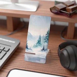 May include: A phone case with a watercolor painting of a snowy mountain landscape with trees. The phone case is on a wooden surface with a keyboard and headphones.