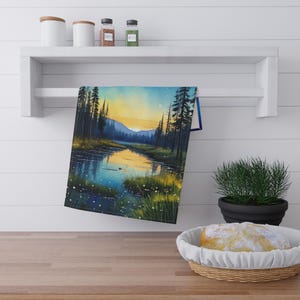 May include: A watercolor painting of a lake in the woods at sunset. The lake is surrounded by trees and the water is reflecting the orange and yellow colors of the sunset. The painting is on a kitchen towel.