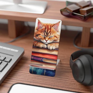 May include: A phone stand featuring a cute orange tabby cat sleeping on a stack of colorful books. The cat has its eyes closed and its paws are resting on the top book. The phone stand is made of a sturdy material and has a sleek design.