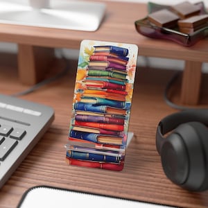 May include: A colorful phone stand with a stack of books printed on it. The stand is made of clear acrylic and has a metal base.