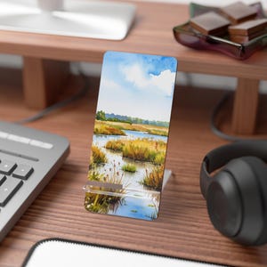 May include: A phone stand featuring a watercolor painting of a marsh landscape with blue sky, water, and tall grasses. The stand is made of clear acrylic and is on a wooden desk. A keyboard and headphones are also visible.