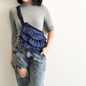 May include: A blue ruffle bum bag with a black strap. The bum bag is worn over a grey long-sleeved shirt and blue jeans.