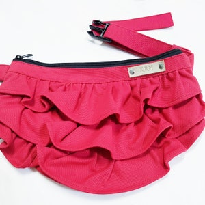 May include: A bright red canvas fanny pack with a black zipper and a black adjustable strap. The fanny pack has three layers of ruffles and a silver metal nameplate with the letters "KRM" on it.