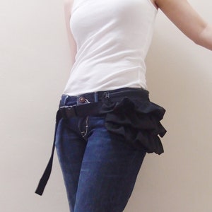 May include: A black ruffle fabric belt with a black adjustable buckle. The belt is worn over a pair of blue jeans.