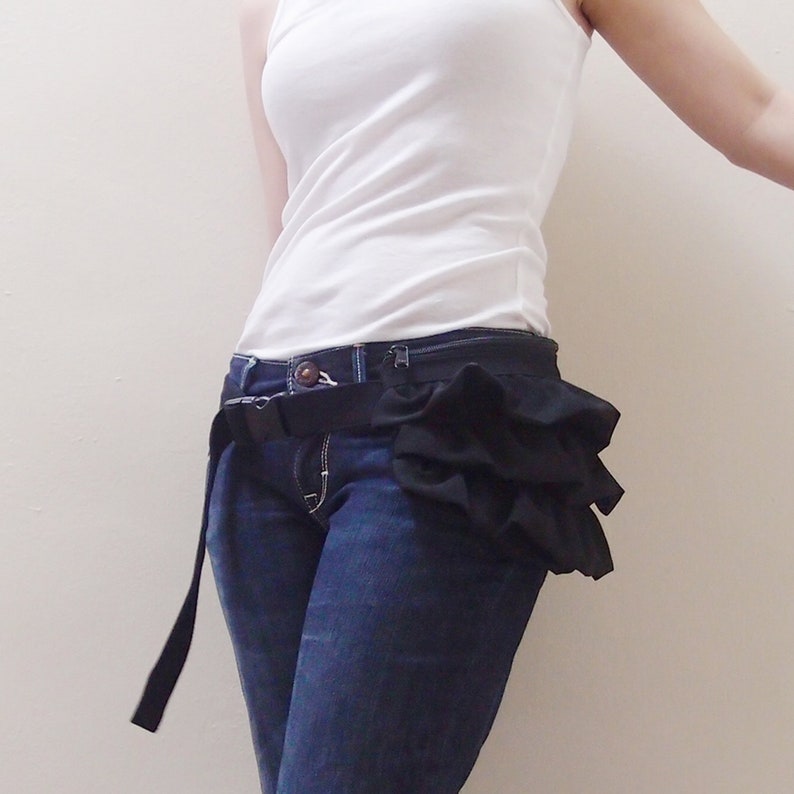 May include: A black ruffle fabric belt with a black adjustable buckle. The belt is worn over a pair of blue jeans.