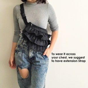 May include: A black ruffle fanny pack worn across the chest. The bag has a zipper closure and is worn with a gray long-sleeve shirt and blue jeans.