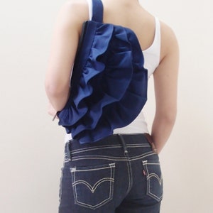 May include: A blue ruffled fabric bag worn as a crossbody bag. The bag is worn over a white tank top and blue jeans.