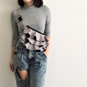 May include: A gray fanny pack with a layered ruffle design. The bag has a black zipper and strap, worn across the body. The model is wearing a gray long-sleeve shirt and acid-wash jeans with a heart-shaped cutout.