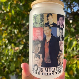Op de afbeelding: Een matglazen beker met een bamboe deksel. De beker heeft een collage van afbeeldingen van een man, met de tekst "KLAUS MIKAELSON THE ERAS TOUR" op de voorkant. De achtergrond is wazig groen.