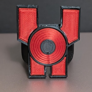 May include: A 3D printed emblem featuring a black frame with a central red circular element and two red rectangular sections at the top and bottom. The design has a layered texture with a black circular center and a black outer border.