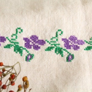 May include: White linen fabric with a green and purple embroidered floral border. The flowers are simple and have five petals.