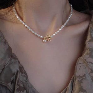 Pode incluir: Um colar delicado de pérolas com um fecho de correr dourado. O colar é feito de pequenas pérolas brancas e tem um design simples e elegante.