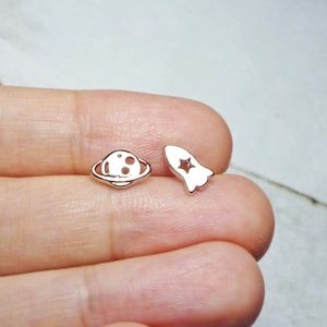 May include: A pair of silver earrings, one shaped like a planet with rings and the other shaped like a rocket with a star inside.