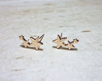 May include: A pair of gold-toned star stud earrings. Each earring features three stars in a row, with the largest star in the center.