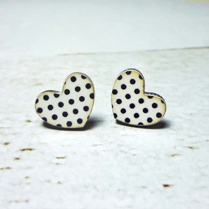 Puede incluir: Un par de pendientes de bot&oacute;n en forma de coraz&oacute;n. Cada pendiente presenta una superficie blanca con lunares negros. Los bordes de los pendientes tienen un borde marr&oacute;n oscuro. Los pendientes se muestran sobre una superficie blanca texturizada.