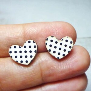 Puede incluir: Un par de pendientes en forma de coraz&oacute;n. Cada pendiente presenta un fondo blanco con lunares negros espaciados uniformemente. Los pendientes tienen un borde dorado. Los pendientes son peque&ntilde;os y parecen estar hechos de madera o un material similar.