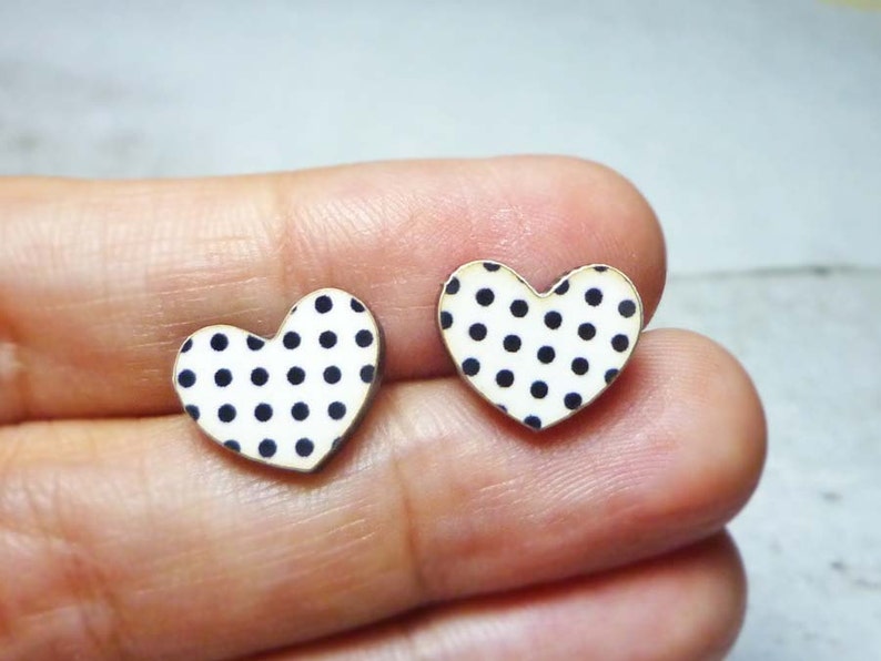 Puede incluir: Un par de pendientes en forma de coraz&oacute;n. Cada pendiente presenta un fondo blanco con lunares negros espaciados uniformemente. Los pendientes tienen un borde dorado. Los pendientes son peque&ntilde;os y parecen estar hechos de madera o un material similar.