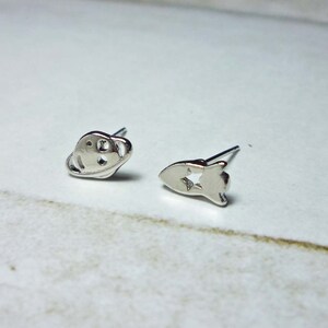 May include: A pair of silver stud earrings, one shaped like a planet with rings and the other shaped like a rocket with a star cutout.