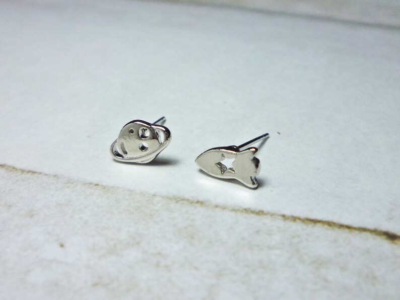 May include: A pair of silver stud earrings, one shaped like a planet with rings and the other shaped like a rocket with a star cutout.