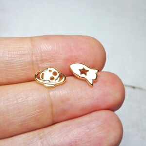 May include: A pair of gold-toned earrings featuring a rocket and a planet design. The rocket has a star cut-out and the planet has a ring around it.